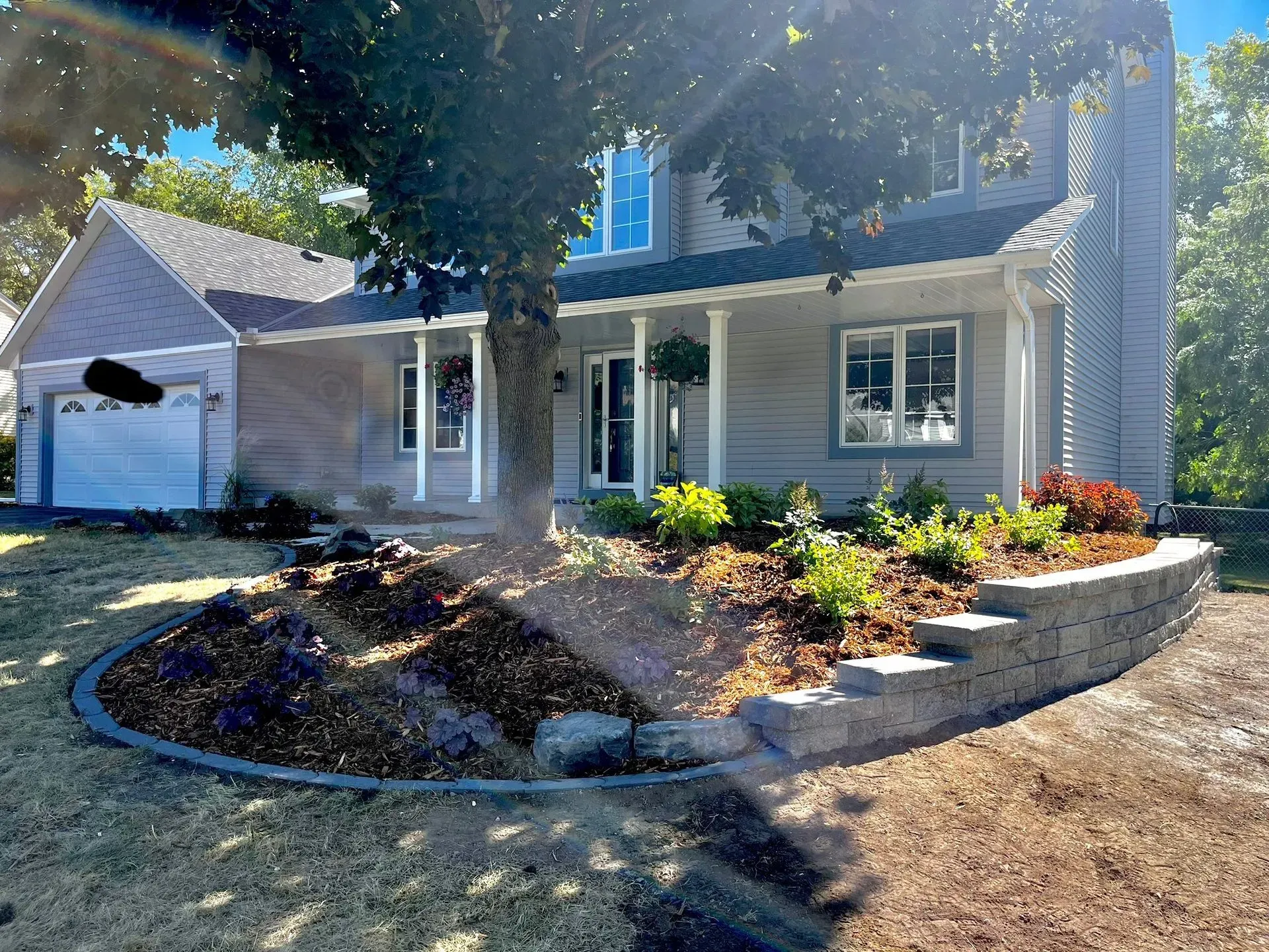 A light gray house with a landscaped front yard featuring a retaining wall and trees on a sunny day.