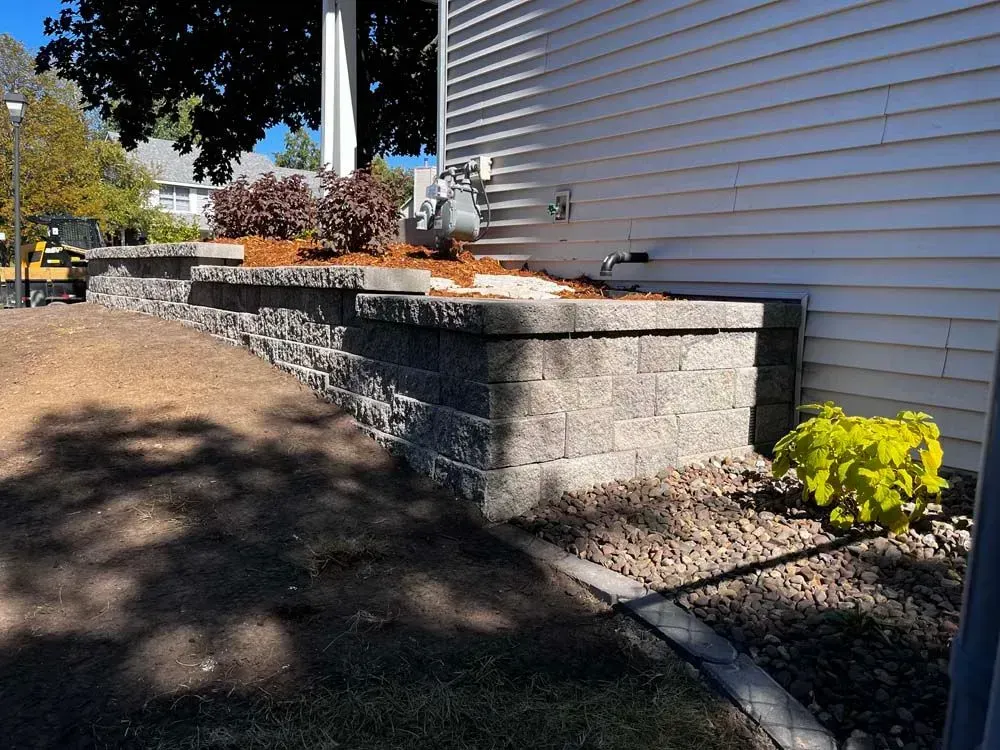 Stone retaining wall with plants and mulch along the side of a building. Brown soil and a curb border the scene.