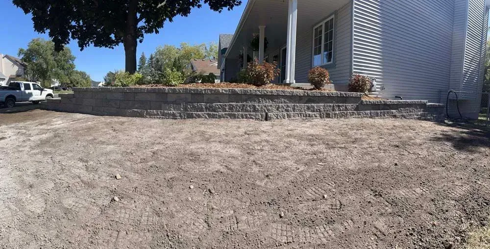 A house with a retaining wall, with exposed dirt in the foreground. The sky is blue.