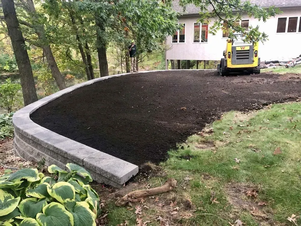 A curved retaining wall supports a freshly tilled area of dark soil, with a small yellow 
