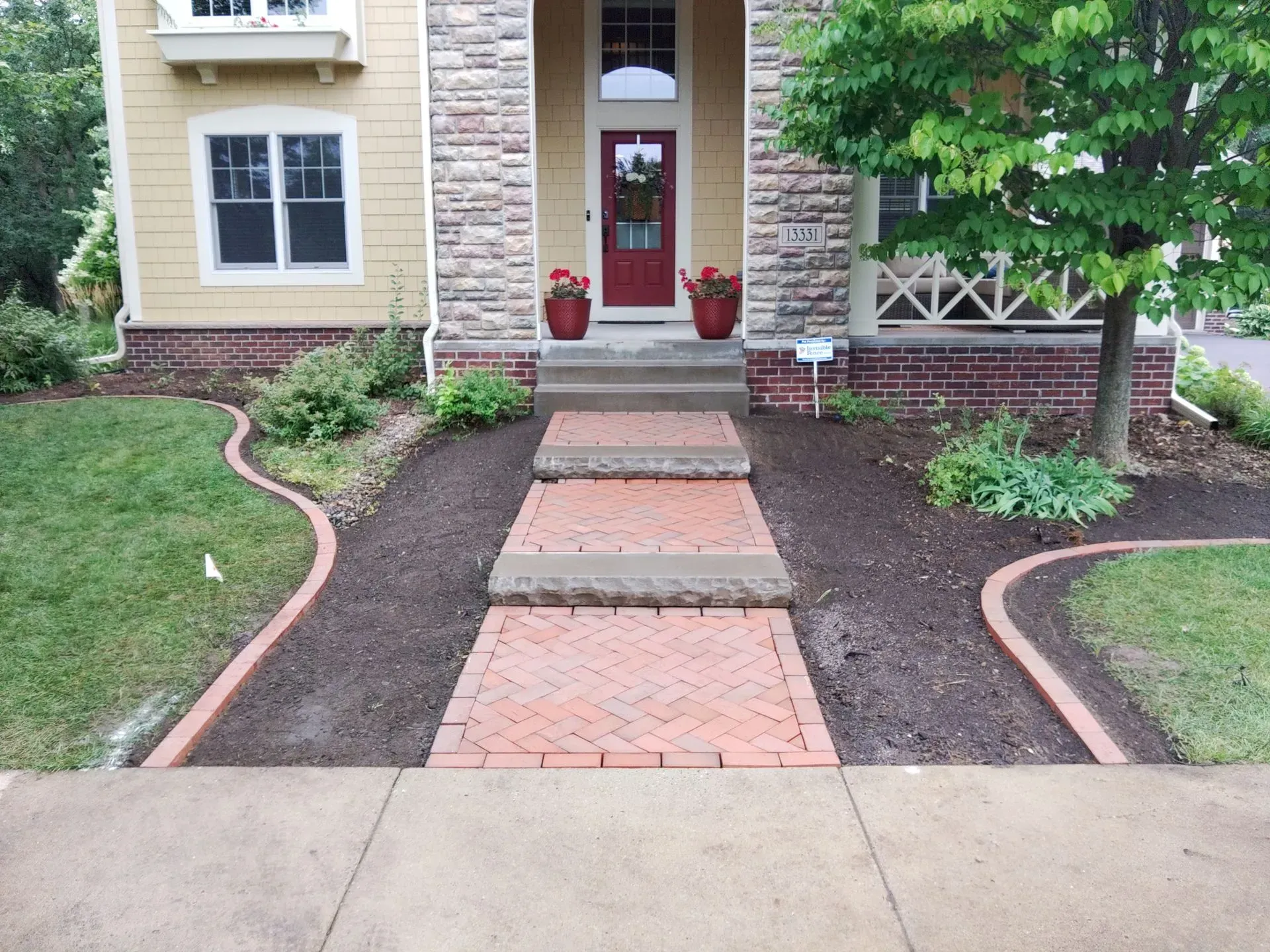 A brick pathway leads to a house with red front door and steps. The pathway is bordered by fresh mulch and green grass.