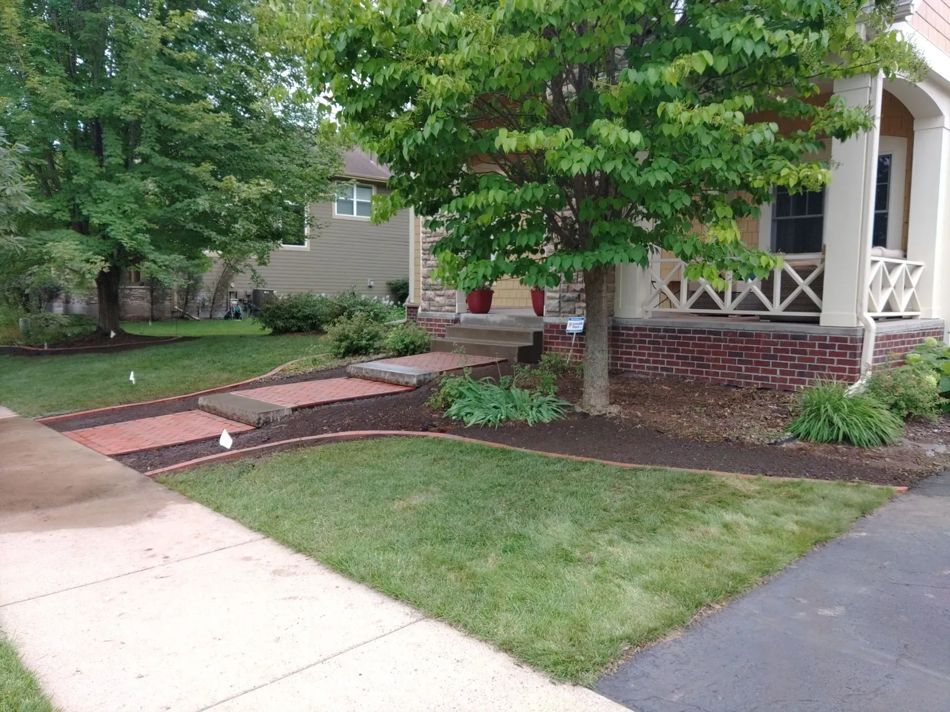 Lush green lawn and red brick steps lead to a house with a porch and mature trees. A sidewalk is in the foreground.