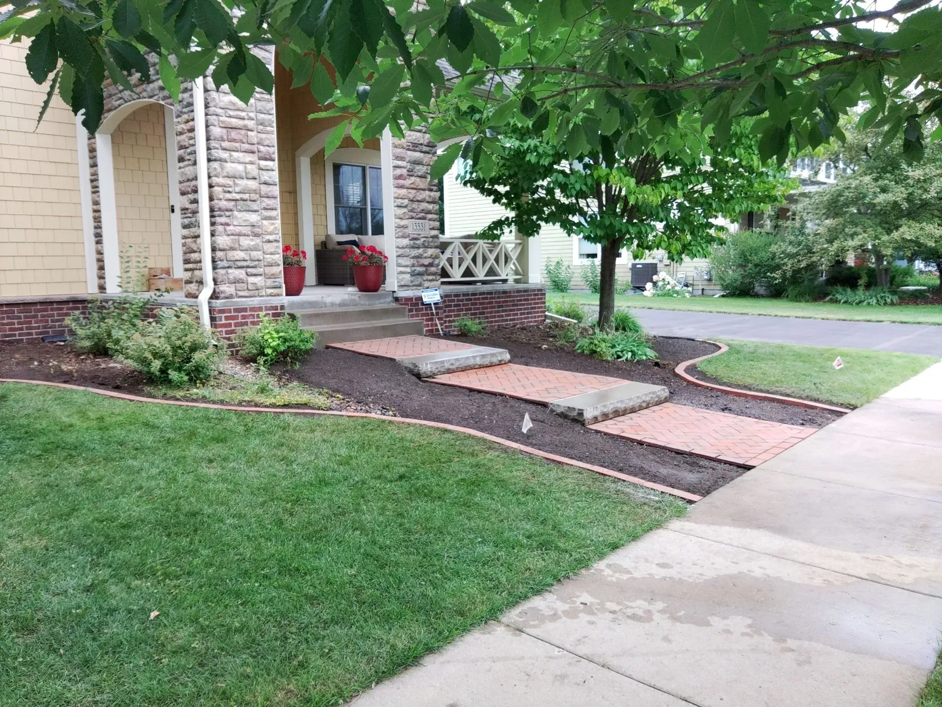 A house exterior with a brick walkway. Green lawn in the foreground, garden beds with mulch and plants.