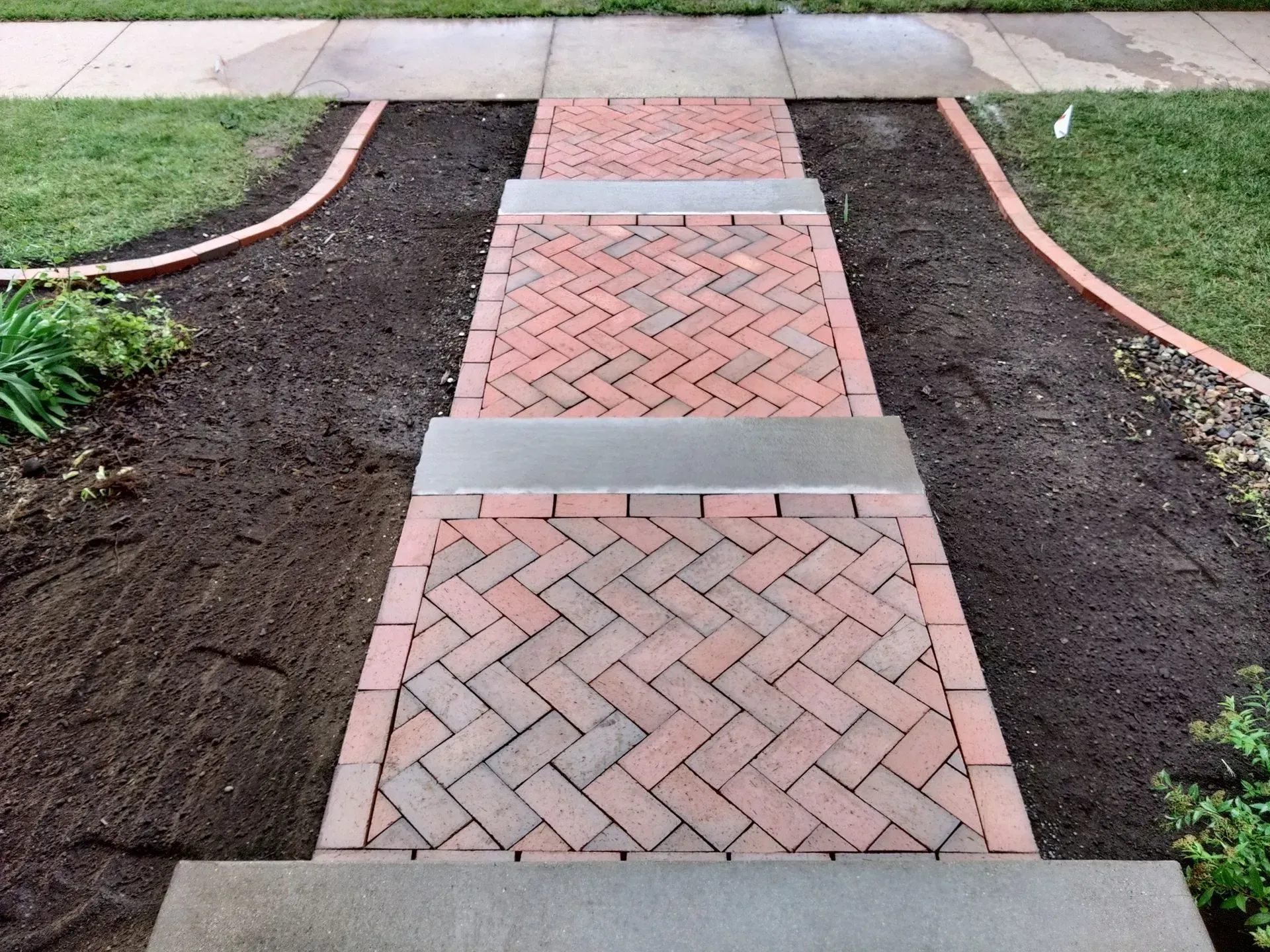 Brick steps leading up to a sidewalk, bordered by freshly tilled dark soil and green grass.