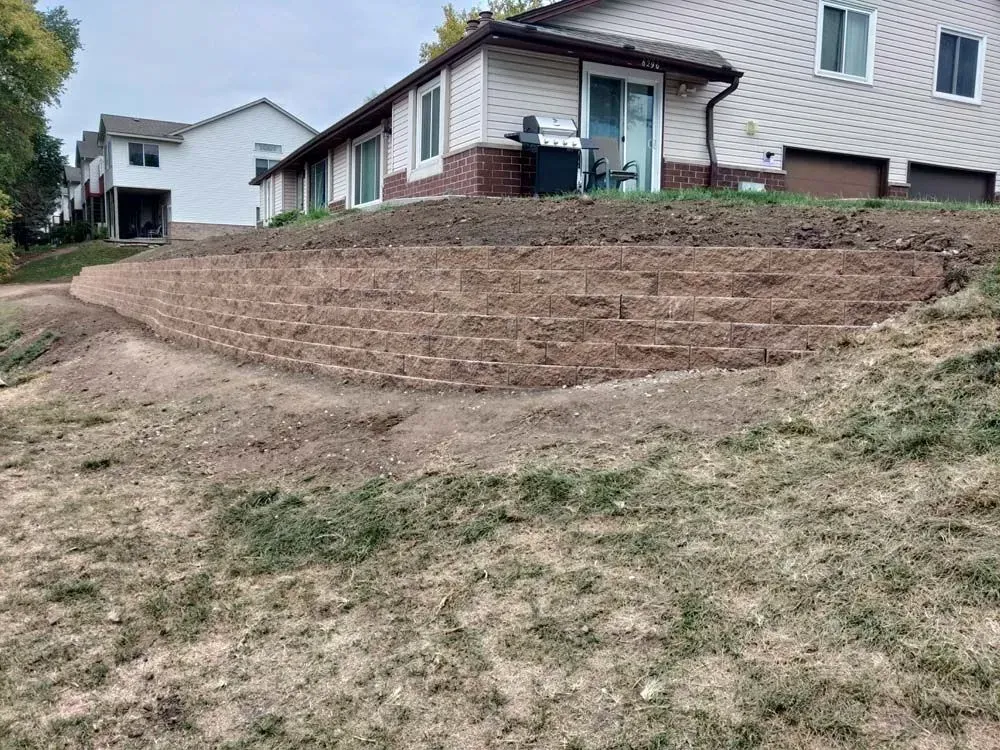 A tiered retaining wall of dirt in front of a house. The house is light-colored with a brown brick base.
