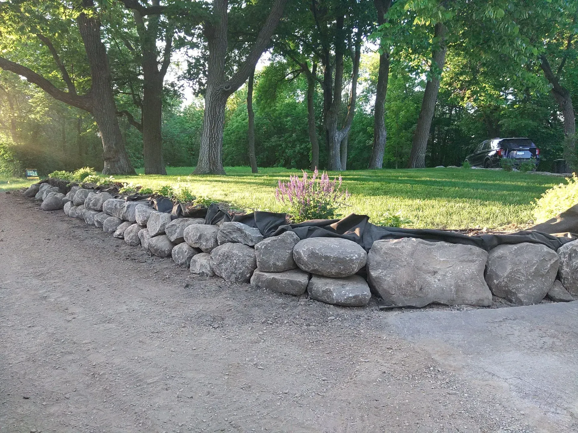 Stone retaining wall along a gravel driveway, with green grass and trees in the background.