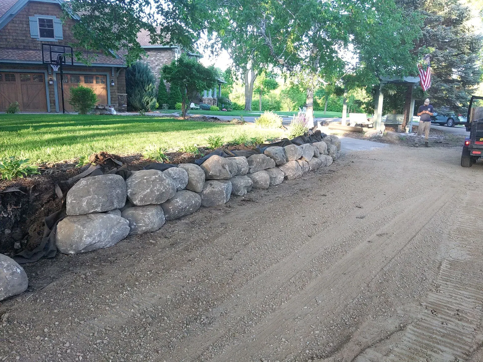 A gravel driveway with a large stone retaining wall on the left. A house and greenery are in the background.