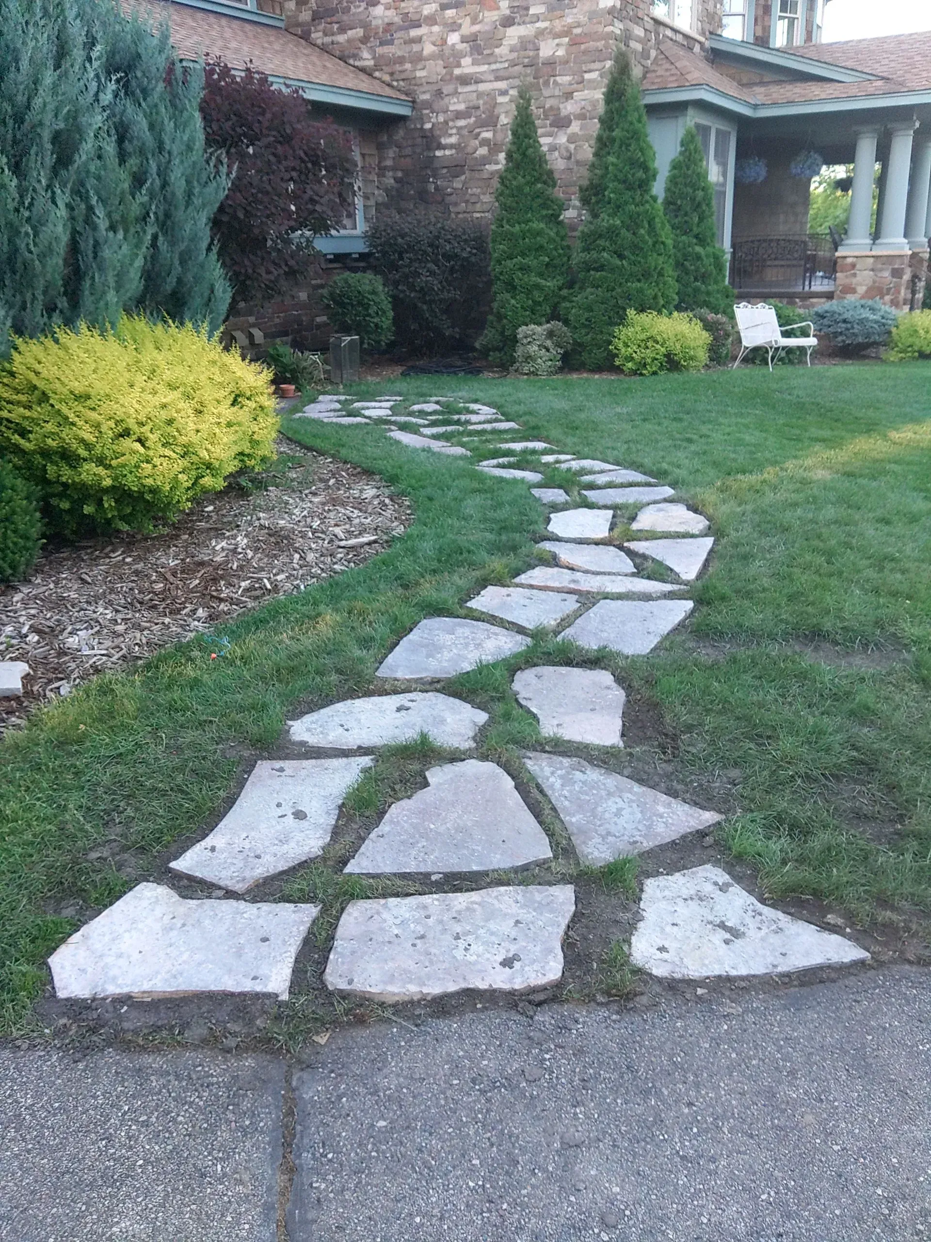 Stone pathway winding through a green lawn towards a house with lush landscaping.