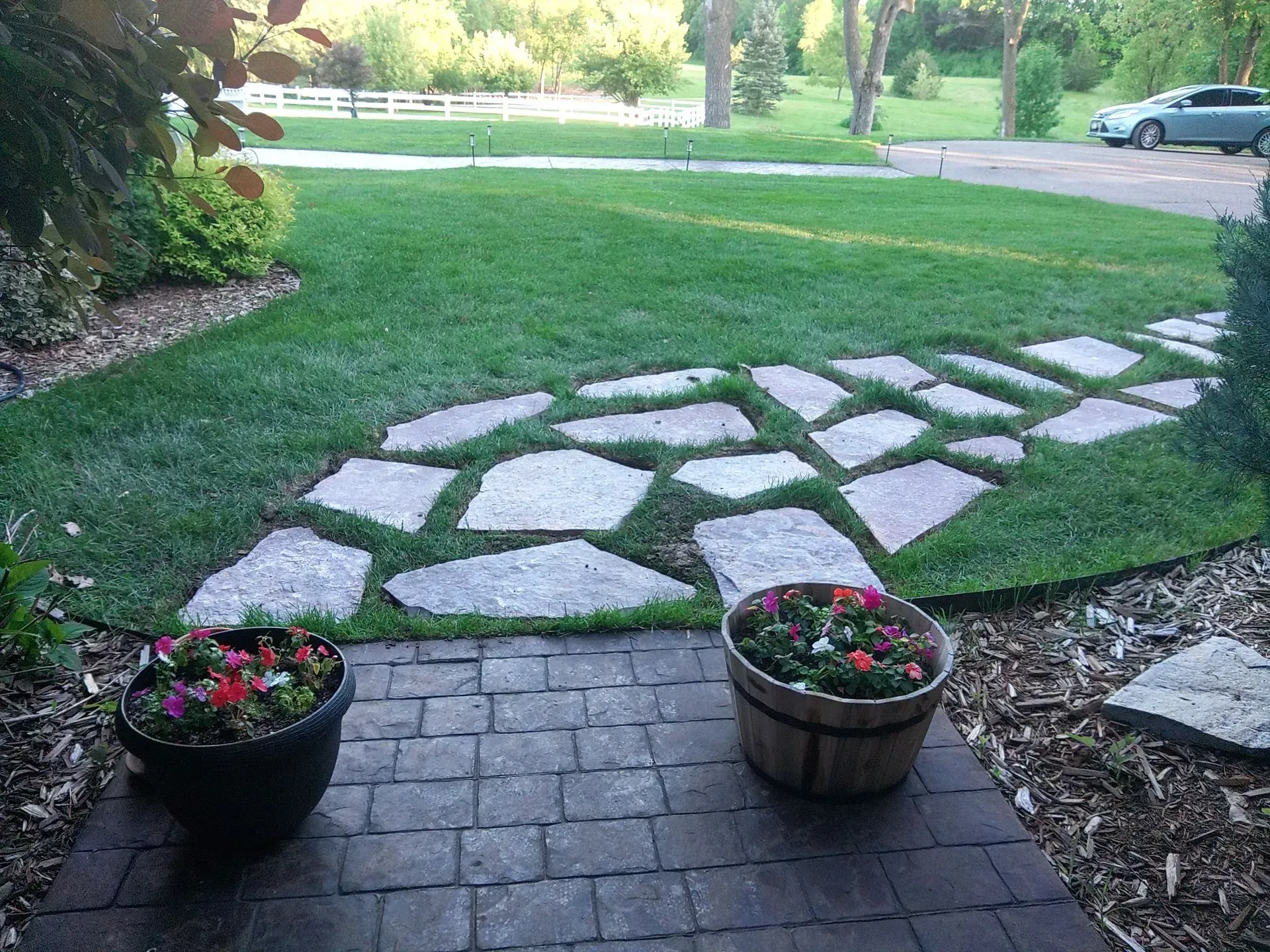 Stone pathway through a green lawn, leading to a yard with trees. Two flower pots flank a brick patio.