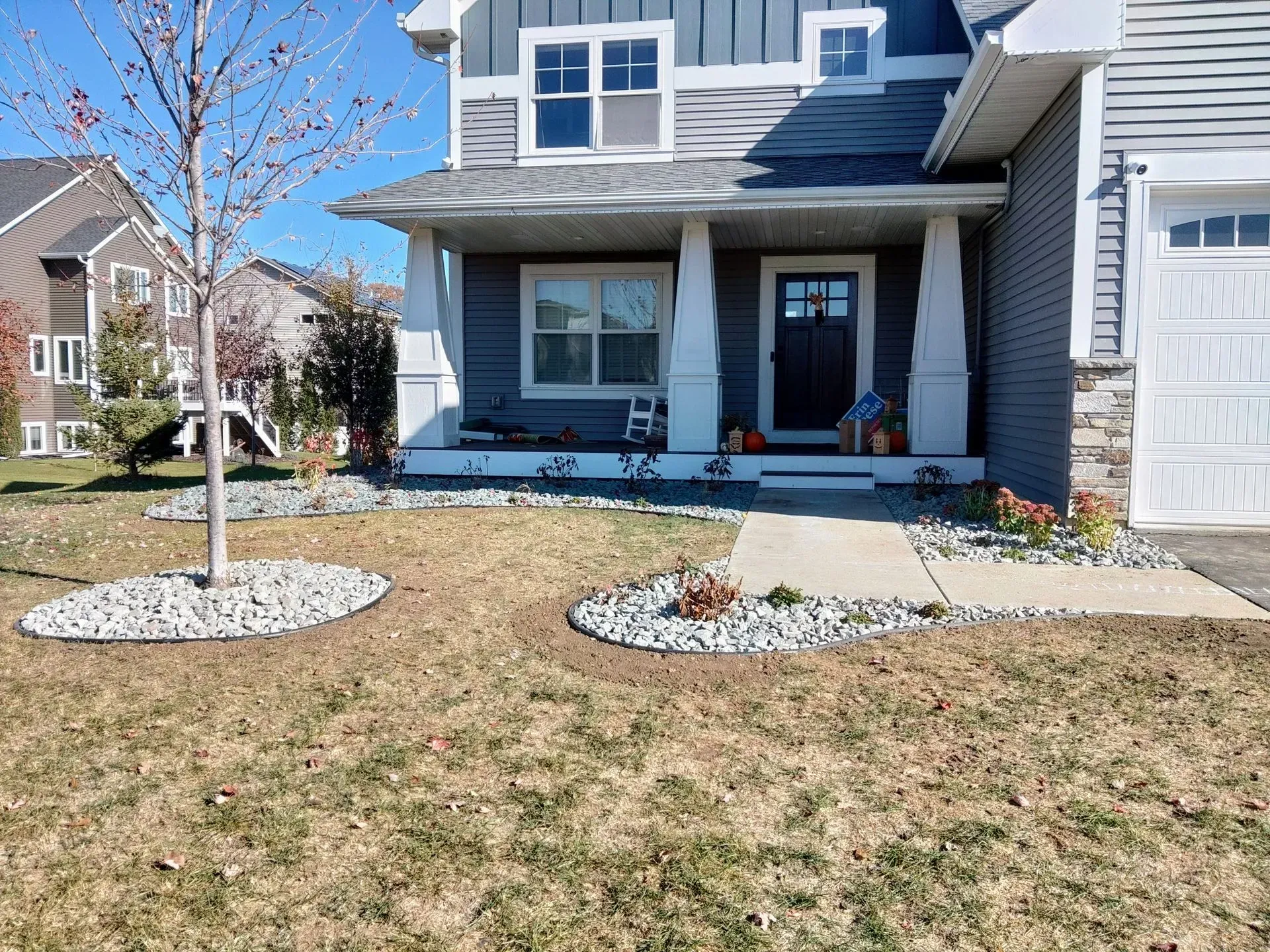 A two-story house with a front porch and a lawn. A small tree is in the yard, surrounded by rocks.