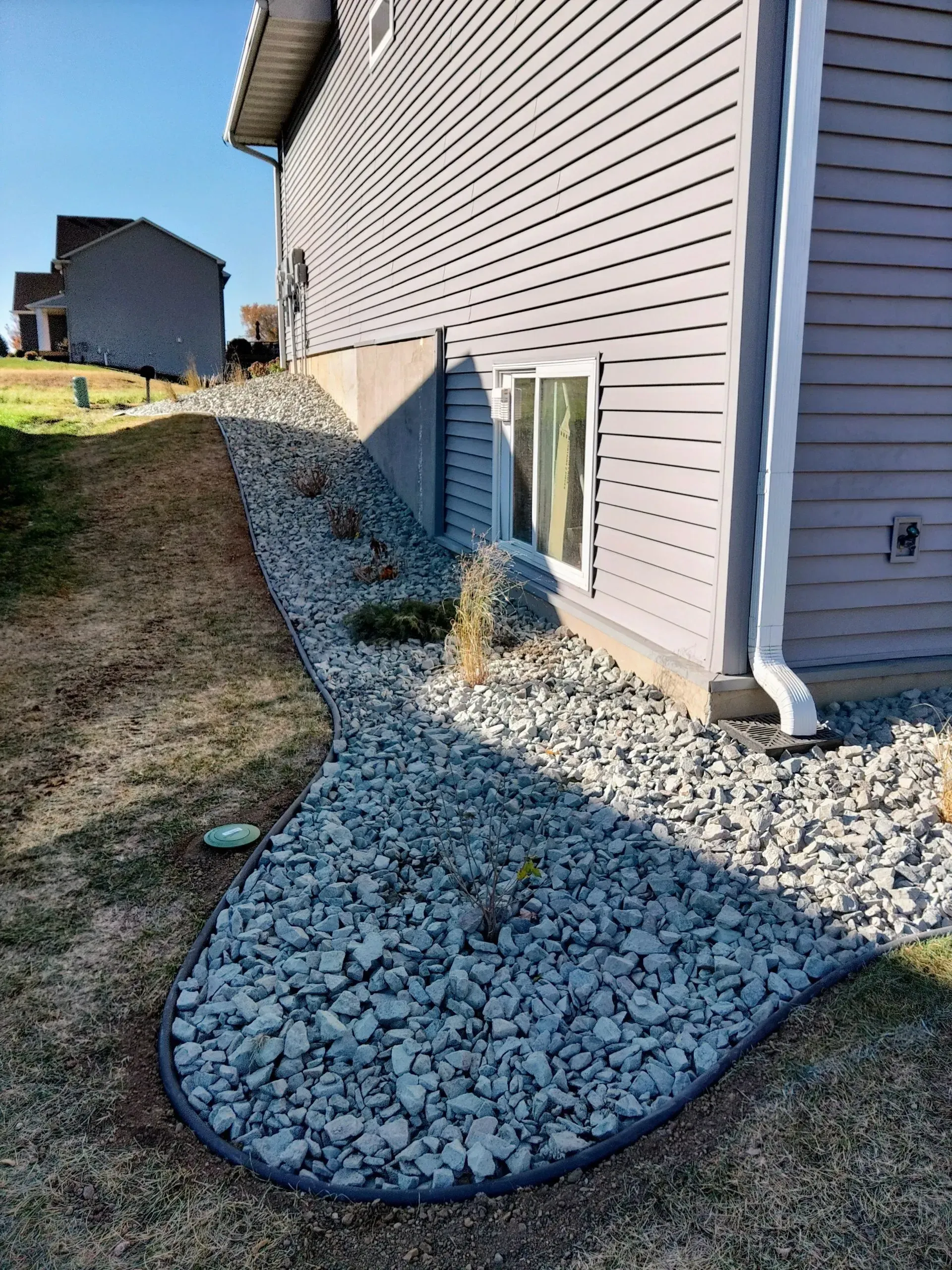 A house exterior with gray siding and a gravel bed along the foundation. The gravel bed borders a grassy slope.