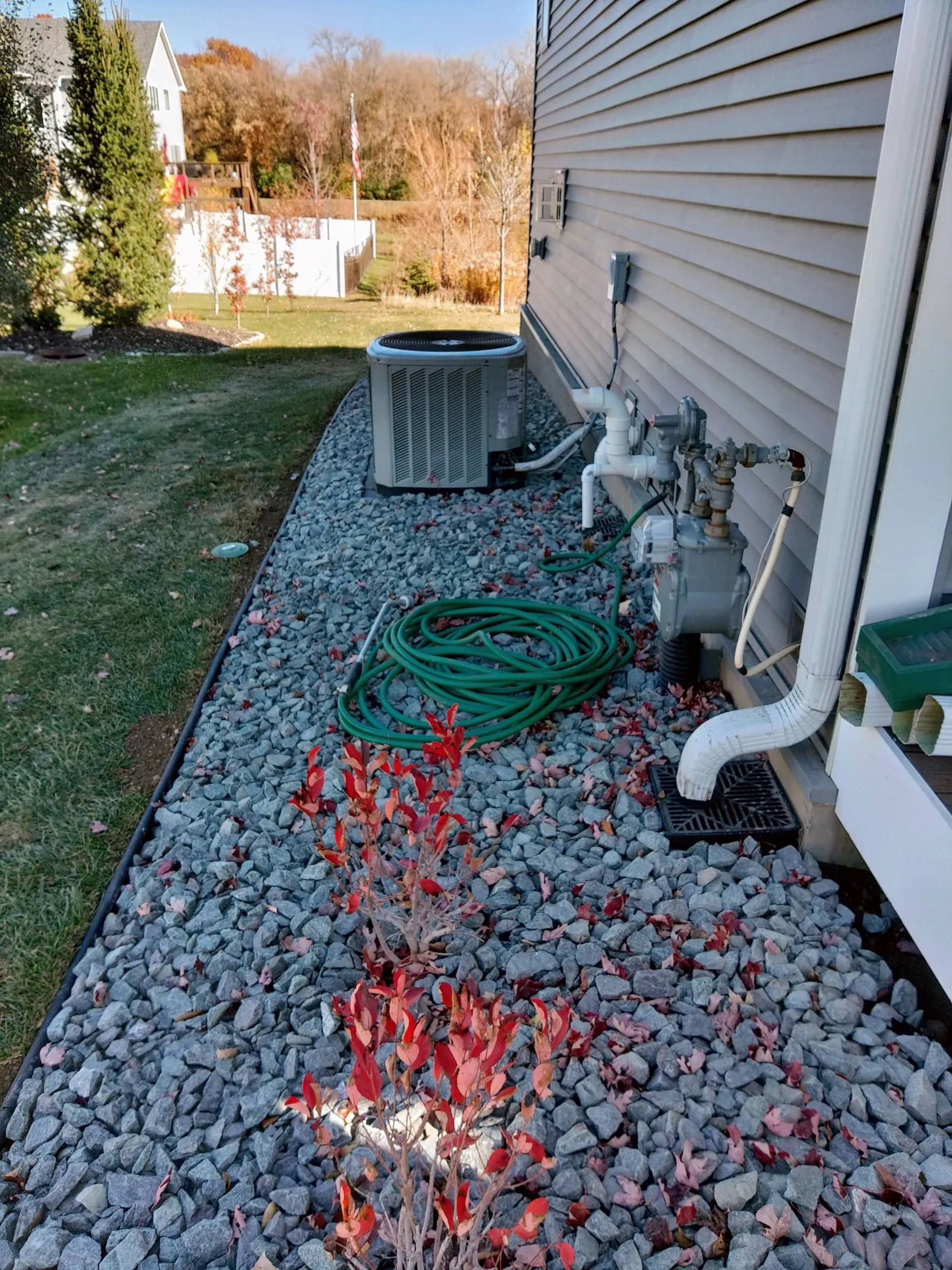 Exterior of a house with an air conditioning unit, water pipes, and a green hose on a bed of gravel.