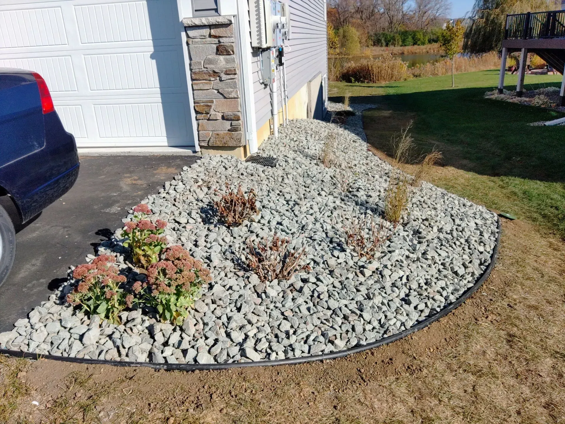 Landscaped corner of a house with gray rocks, edged with black, and some dormant plants. A car is parked nearby.