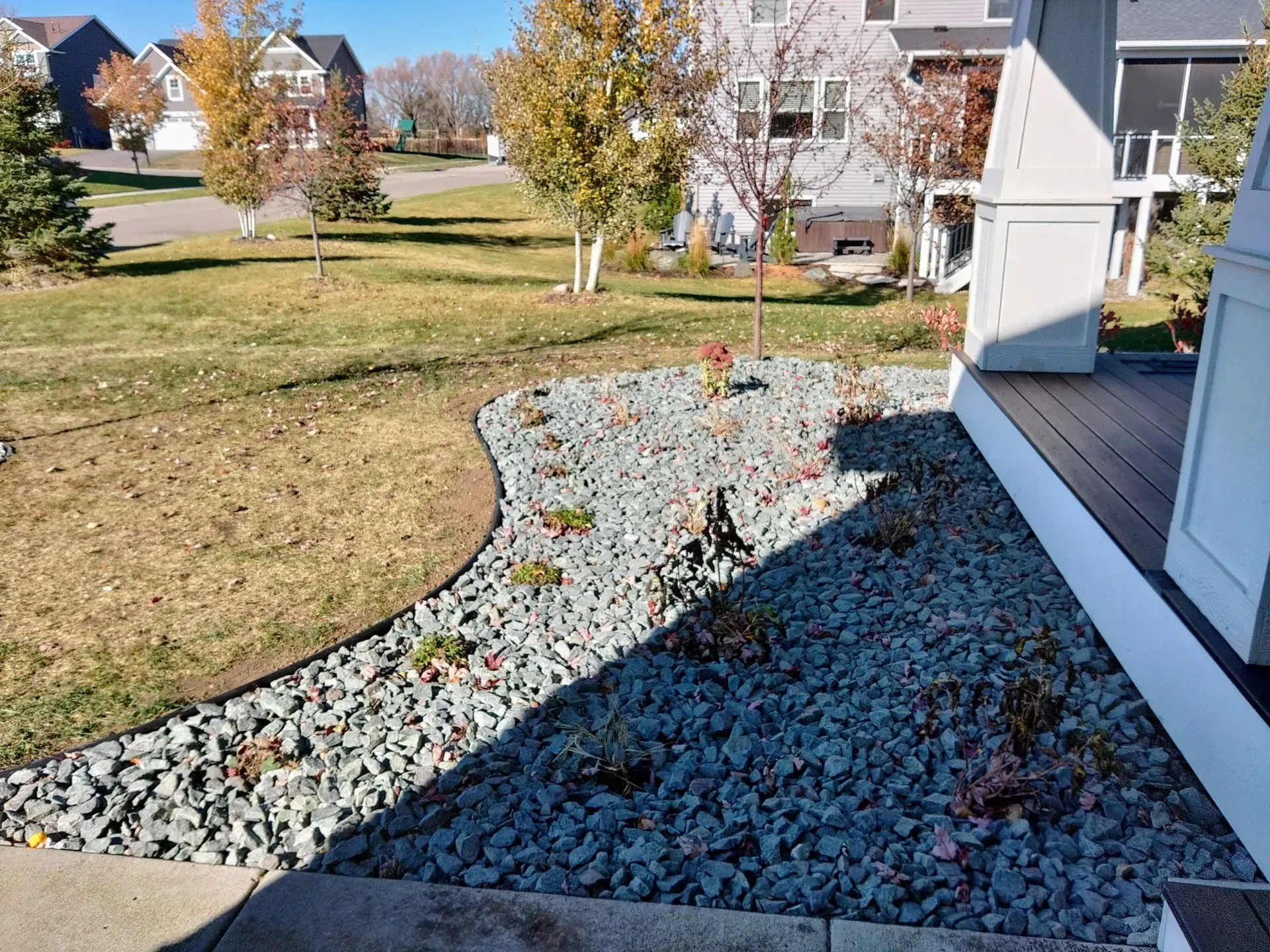 A front yard landscaping with gray river rock, newly planted shrubs, and a brown deck next to a grass lawn.