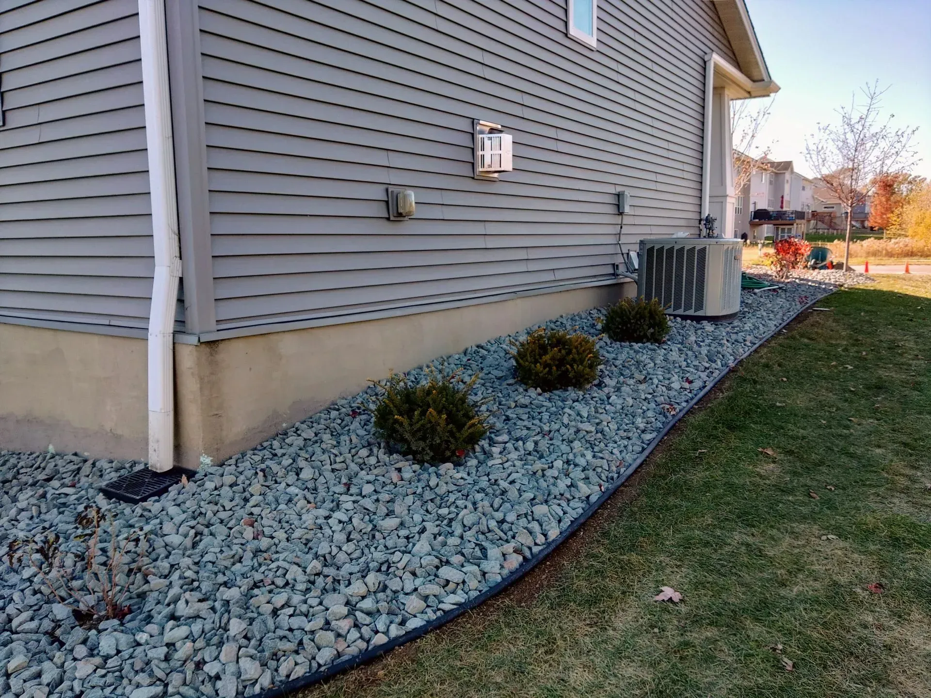 Gray house exterior with blue gravel bed containing three green bushes, adjacent to a grassy lawn.