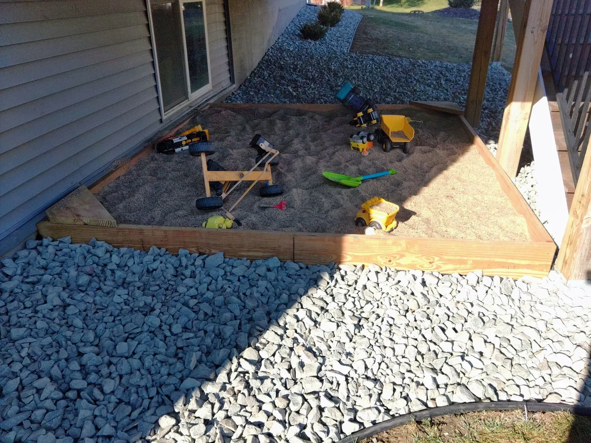 Sandbox with toys, bordered by wooden beams, next to a house and gravel pathway.