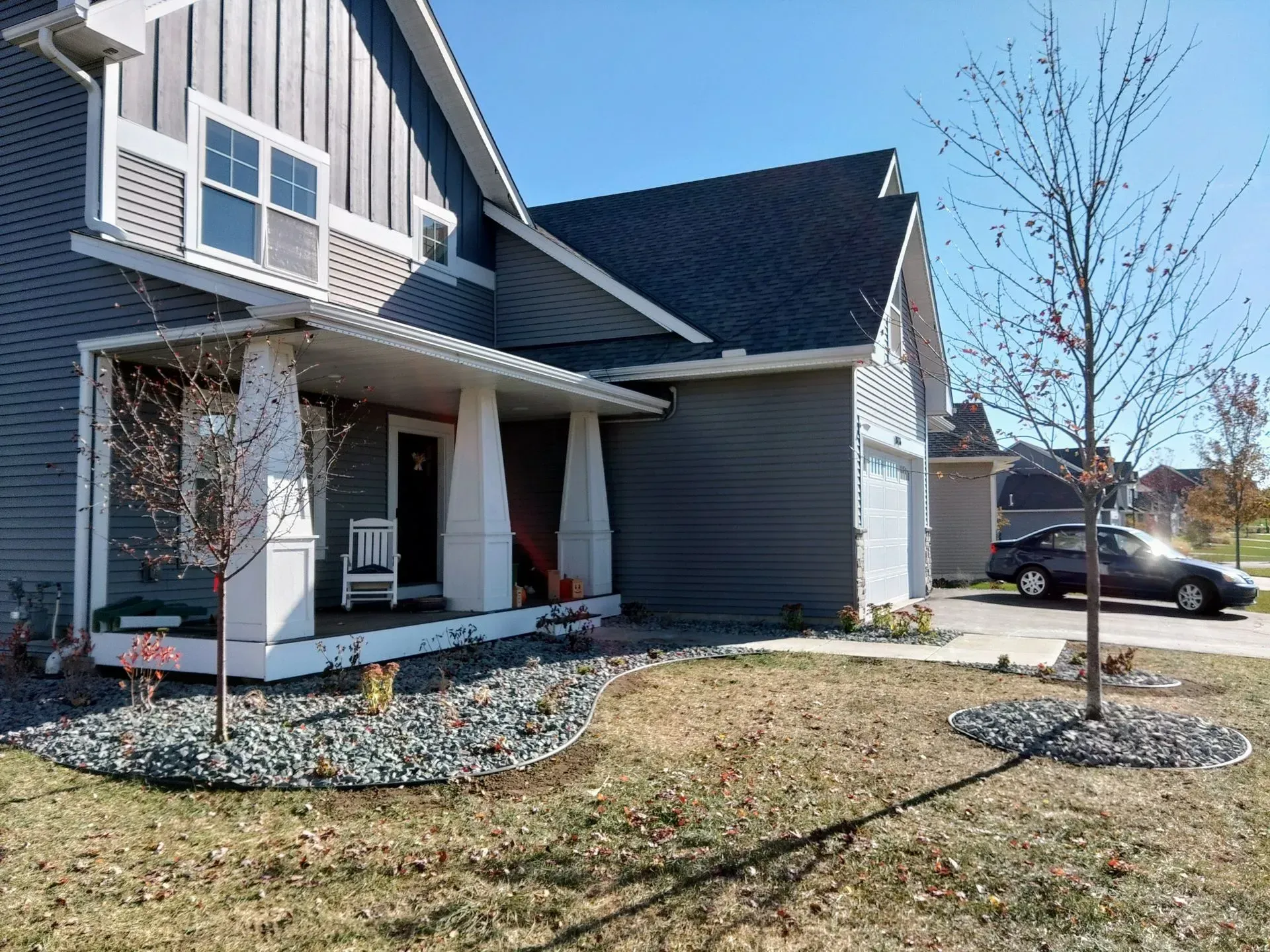 Gray and white house with a porch, a small tree, and a car parked in the driveway on a sunny day.