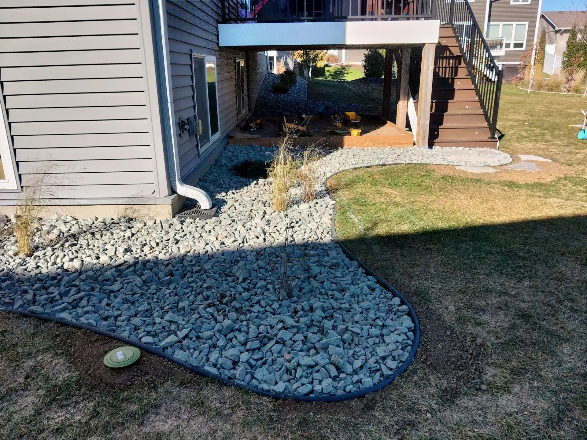 Landscaped backyard with gray rock border against a light gray house. Deck and stairs are visible.