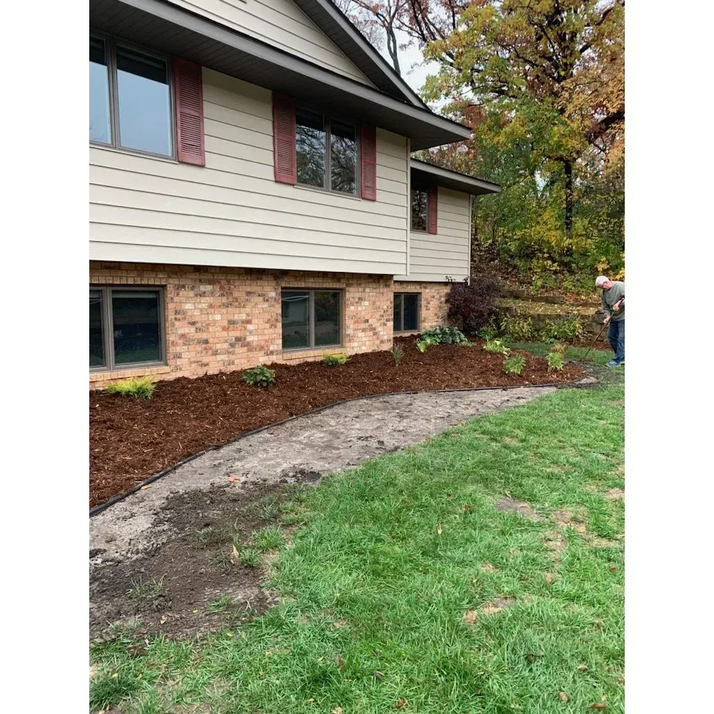 Side view of a two-story house with red shutters and brick, with a newly mulched garden bed in front. 