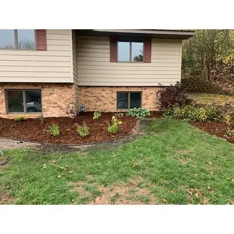 Tan house with brick base and burgundy shutters, surrounded by mulch beds with green plants and lawn.