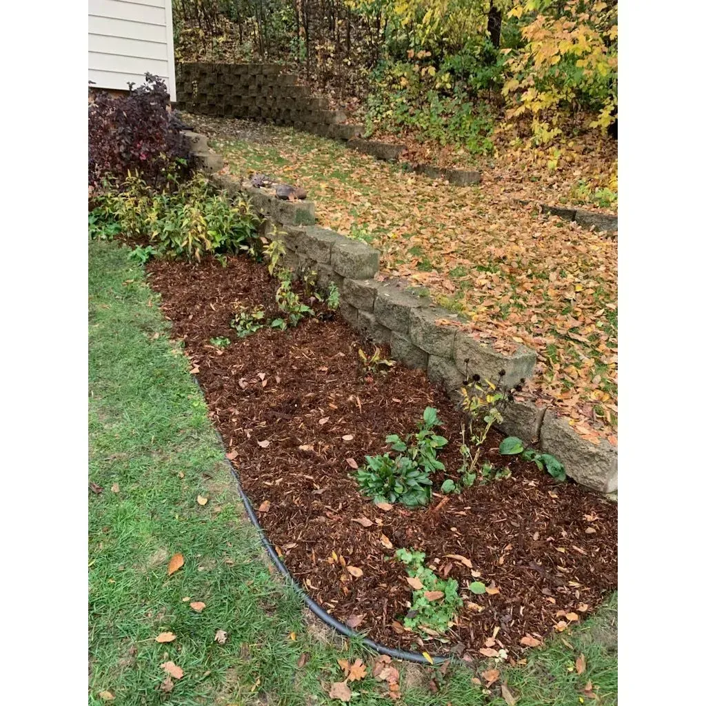 A garden bed with brown mulch and green plants next to a retaining wall, with grass and fallen leaves in the background.