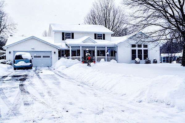 White two-story house covered in snow. A snow-covered driveway with a blue car in the garage.