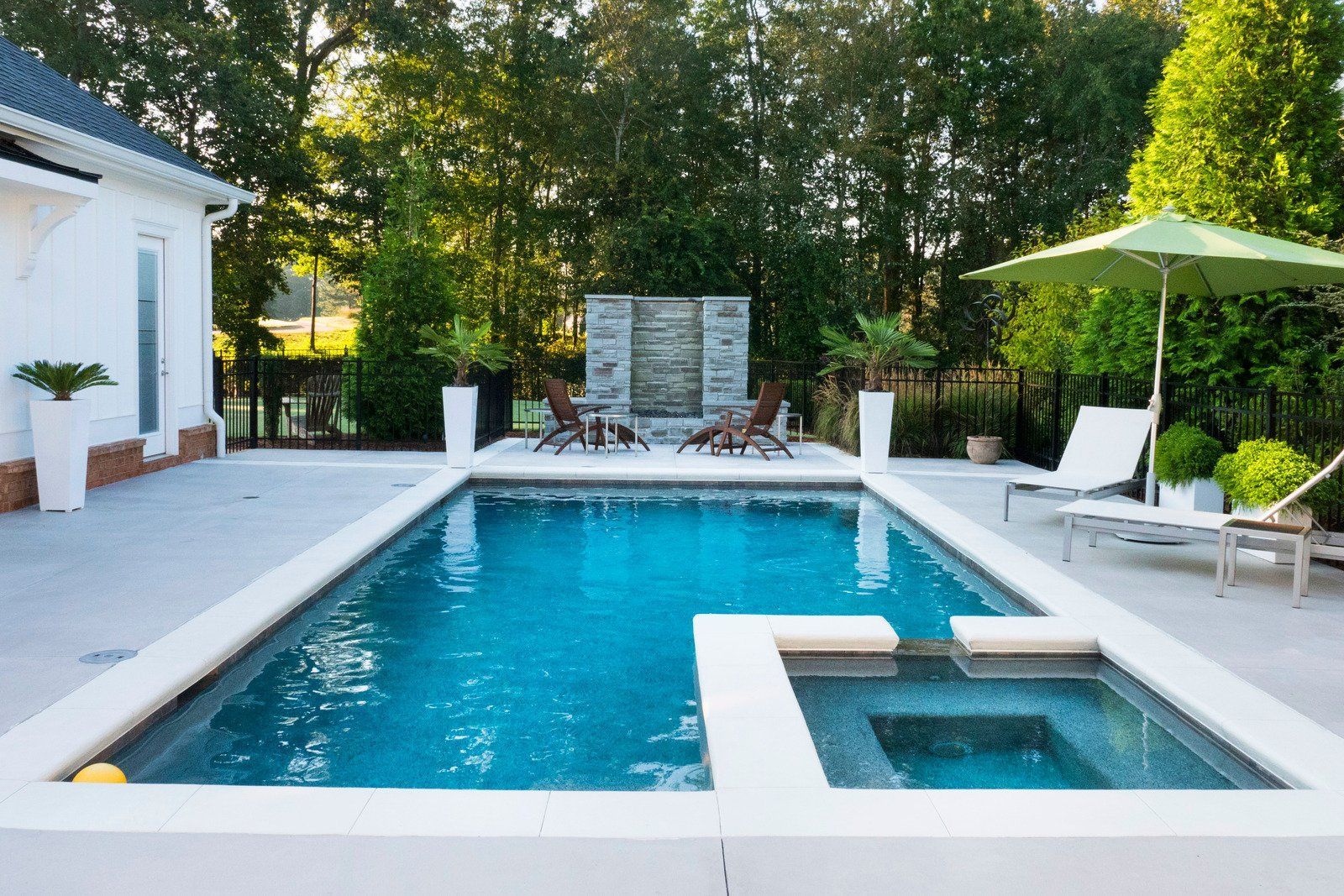 Rectangular pool with a hot tub, patio furniture, and a stone wall waterfall. Green umbrella provides shade.