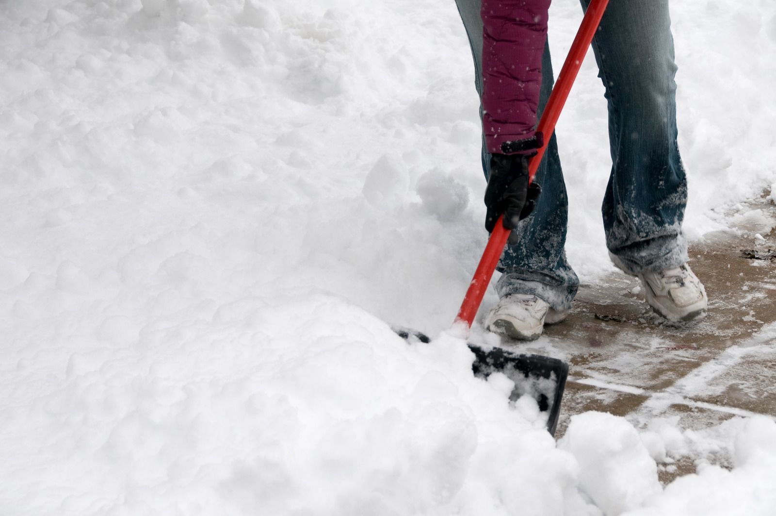 Person shoveling snow from a sidewalk, using a red shovel.