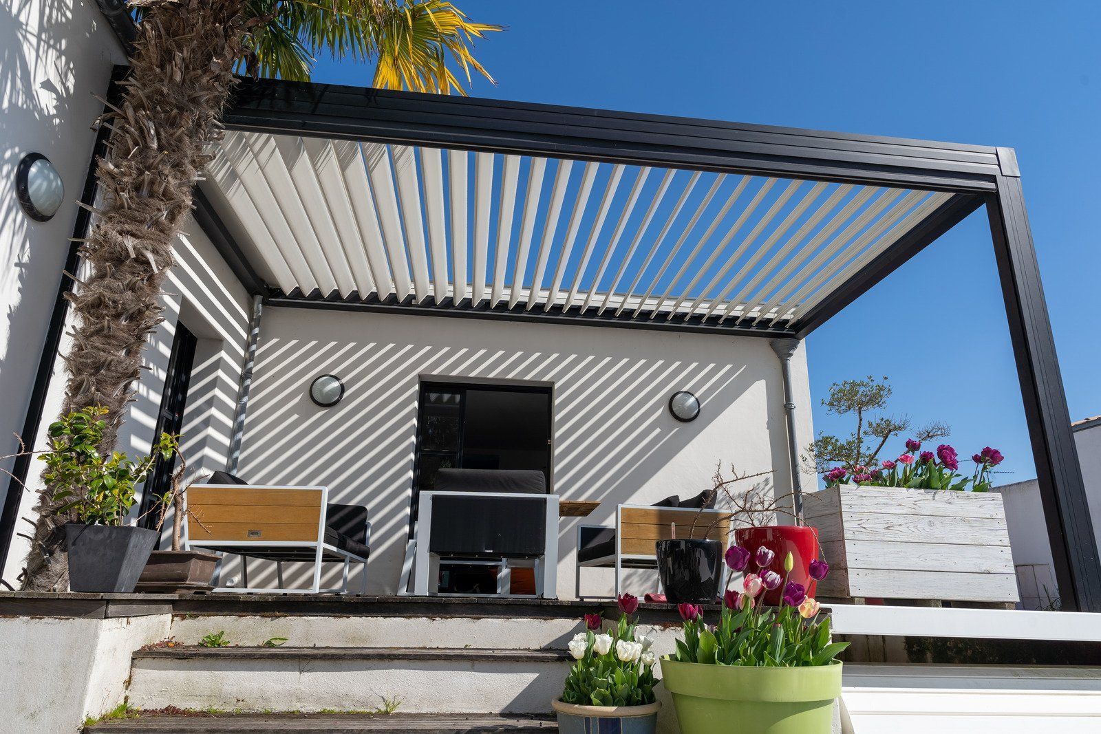 Pergola with adjustable white slats casting shadows on a white-walled patio with seating, plants, and a blue sky.