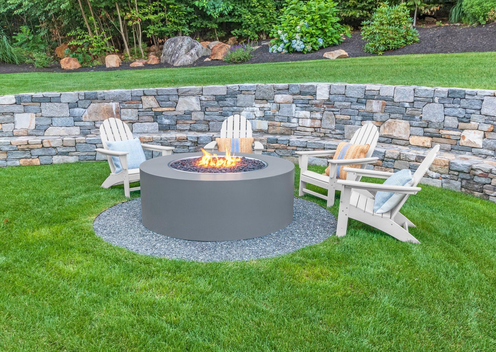 A backyard scene with a gray fire pit, four wooden chairs, and a stone wall. The scene is on a grassy hill.