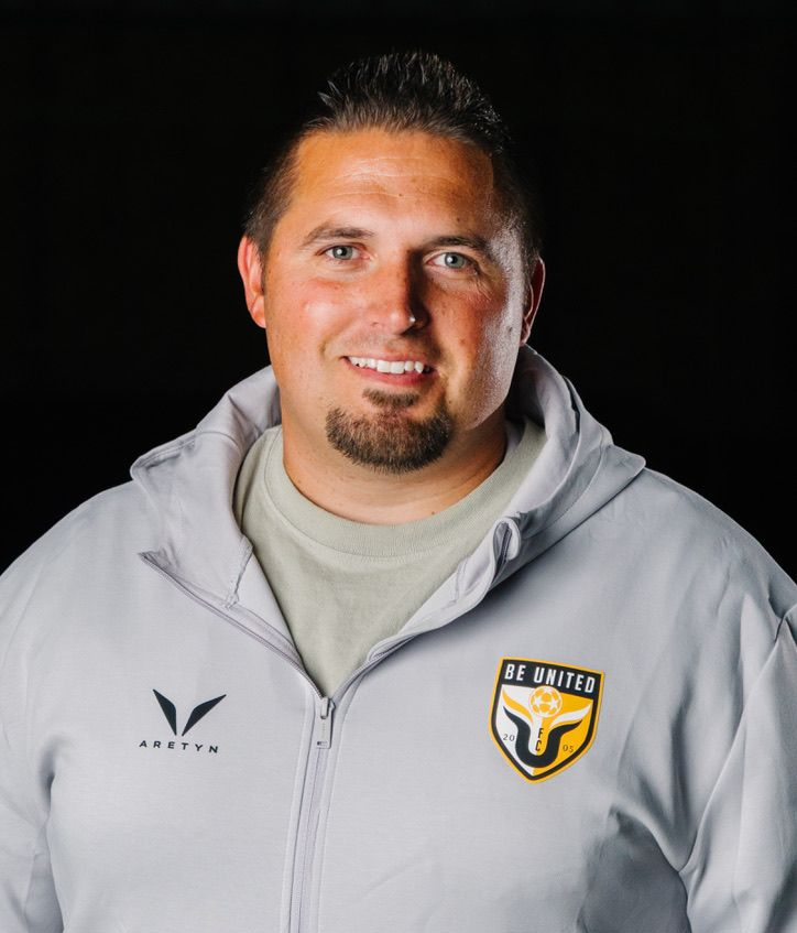 Football coach with arms crossed, looking stern on a sunny field, players in the background.