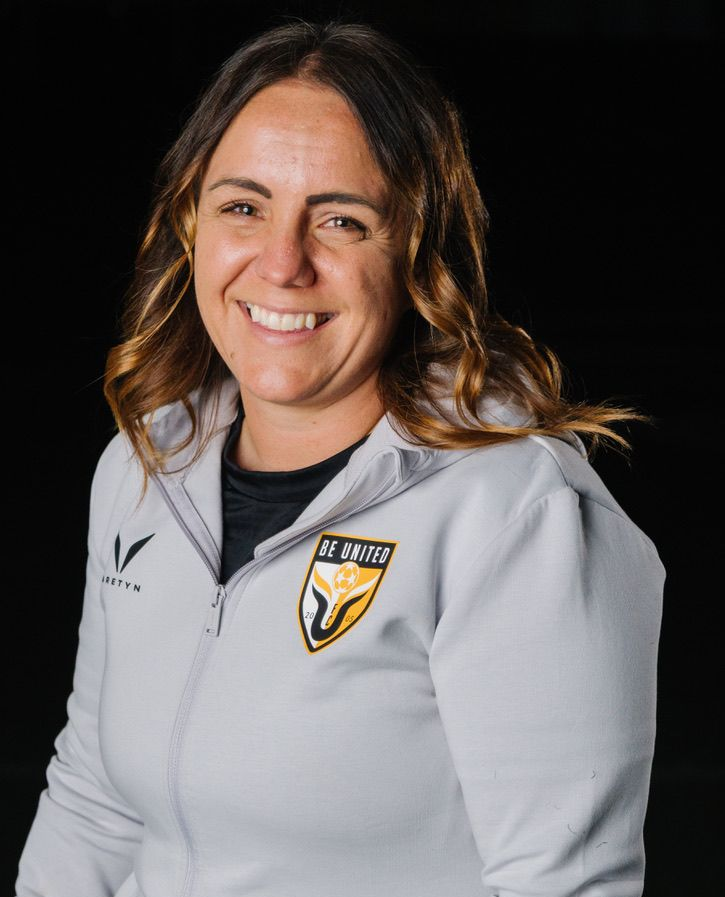 Football coach with arms crossed, standing on a football field. He wears a navy polo shirt and gray pants.
