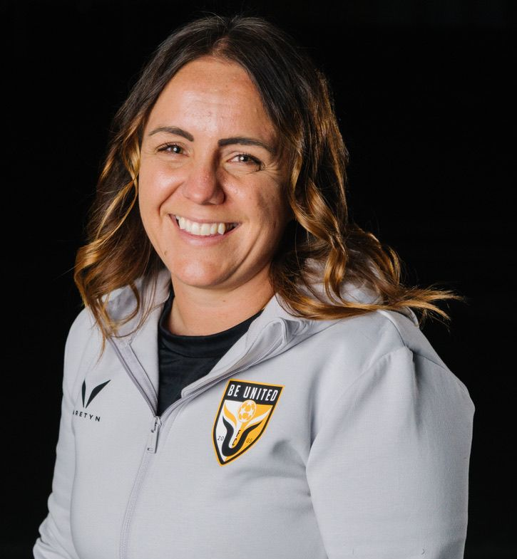 Football coach with arms crossed, standing on a football field. He wears a navy polo shirt and gray pants.