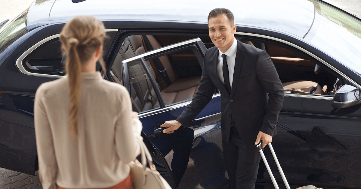 A man in a suit is opening the door of a car for a woman with a suitcase.