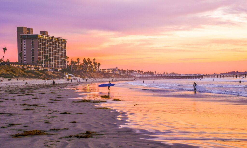 A person is holding a surfboard on the beach at sunset.