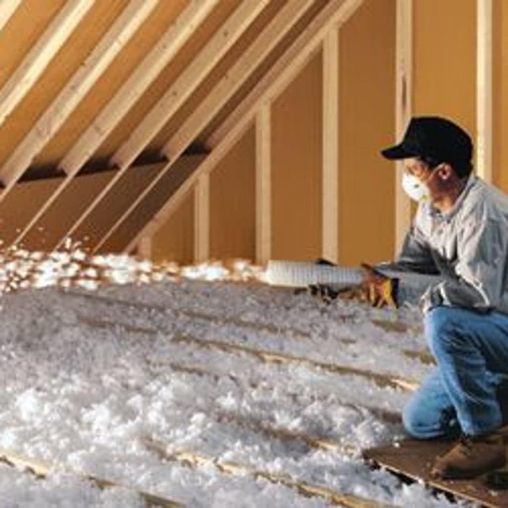 Man in a dusty attic wearing a respirator and gloves, spreading insulation between rafters.