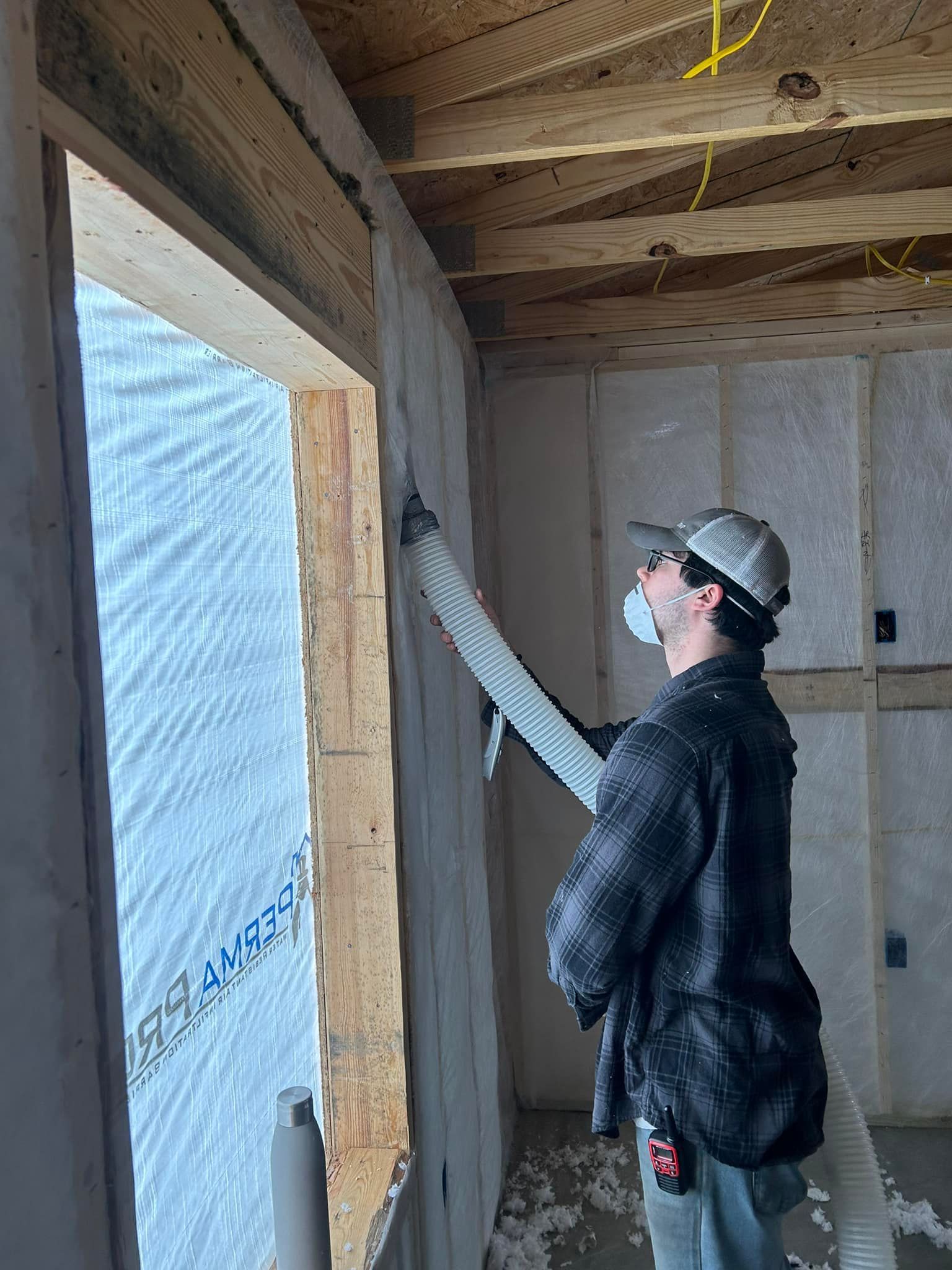 Man insulating a wall near a window. He wears a mask and hat, holding a hose. Interior construction setting.