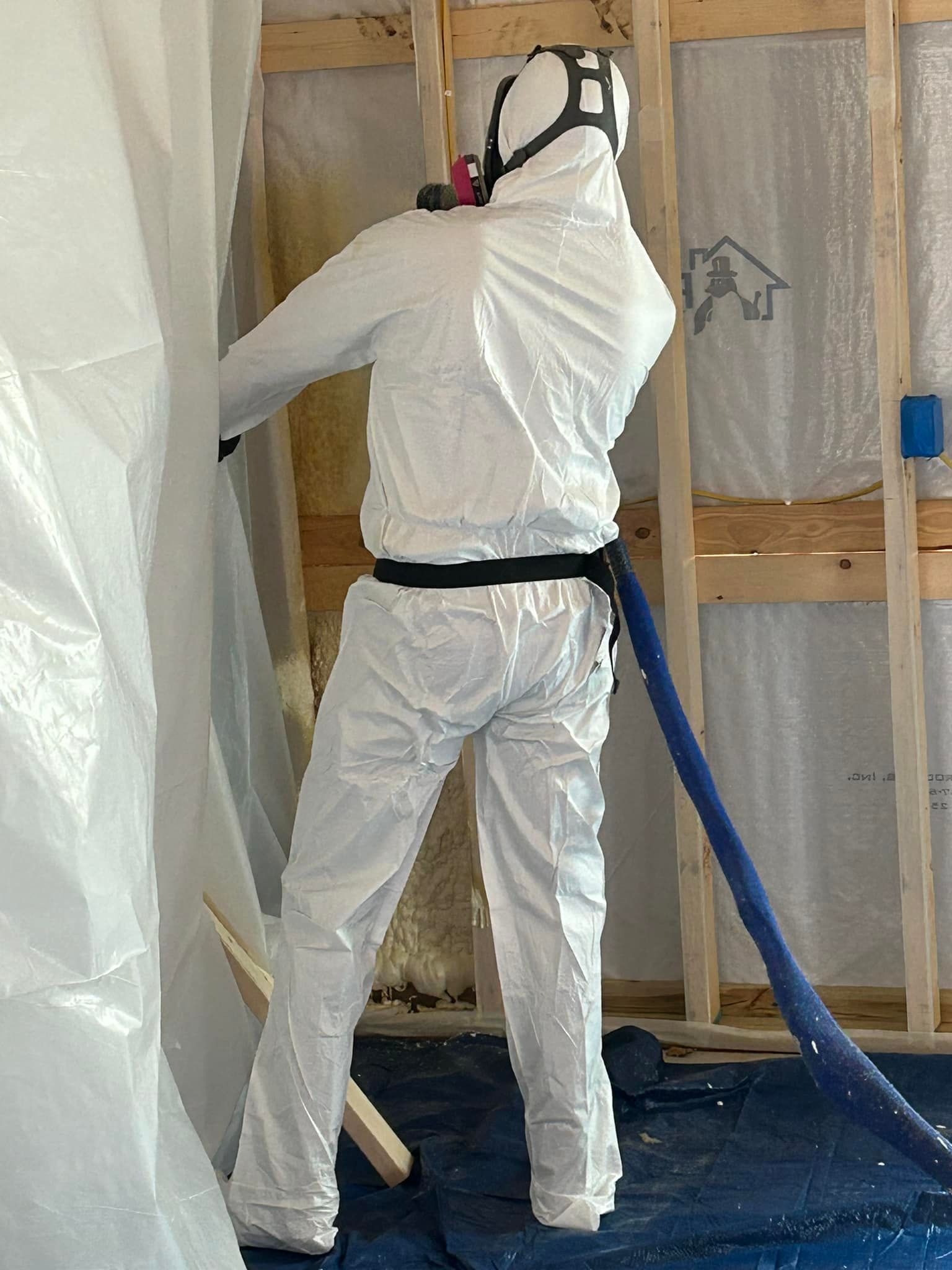 Person in white protective suit spraying insulation inside a wall frame, blue hose attached.