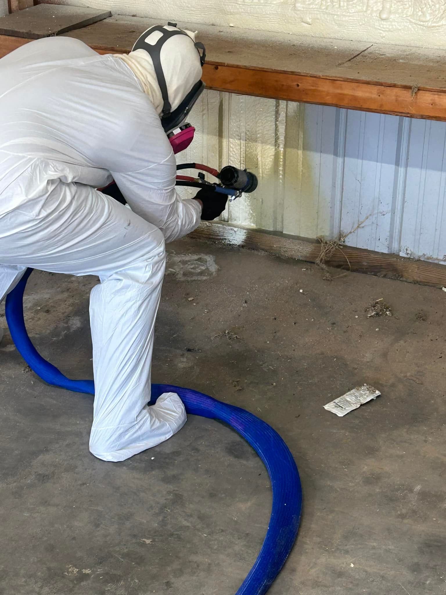 Person in a white protective suit spraying a substance onto a wall. They are holding a nozzle connected to a blue hose.