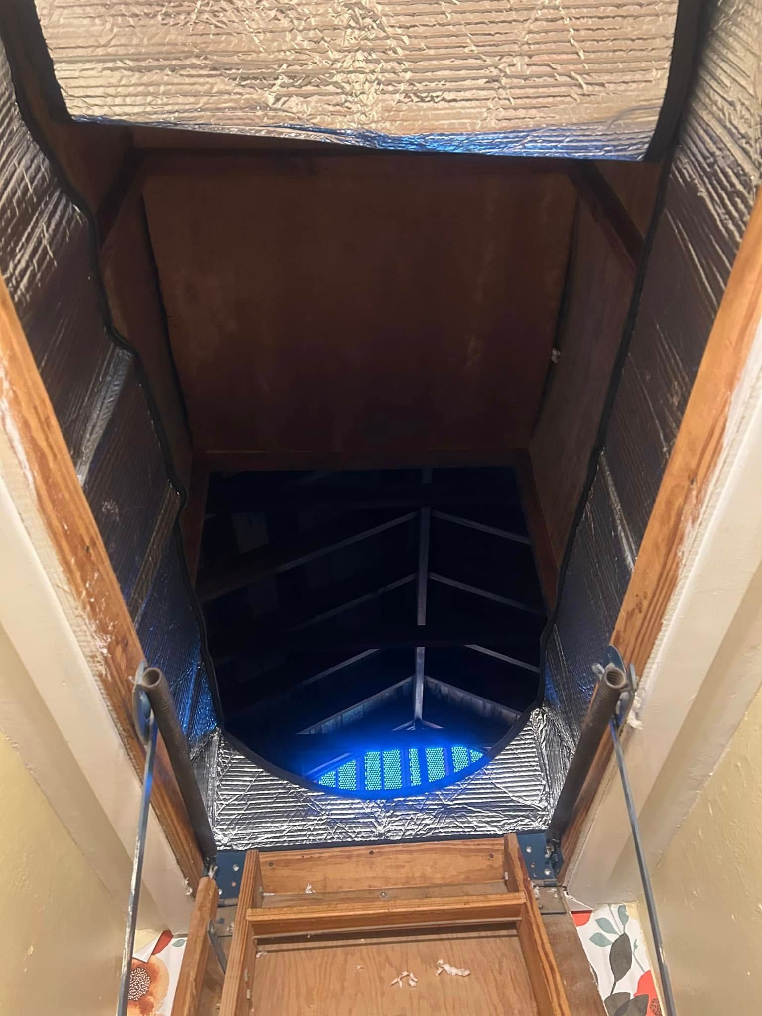 Looking up into an attic access, showing wooden stairs and the dark, insulated opening above.