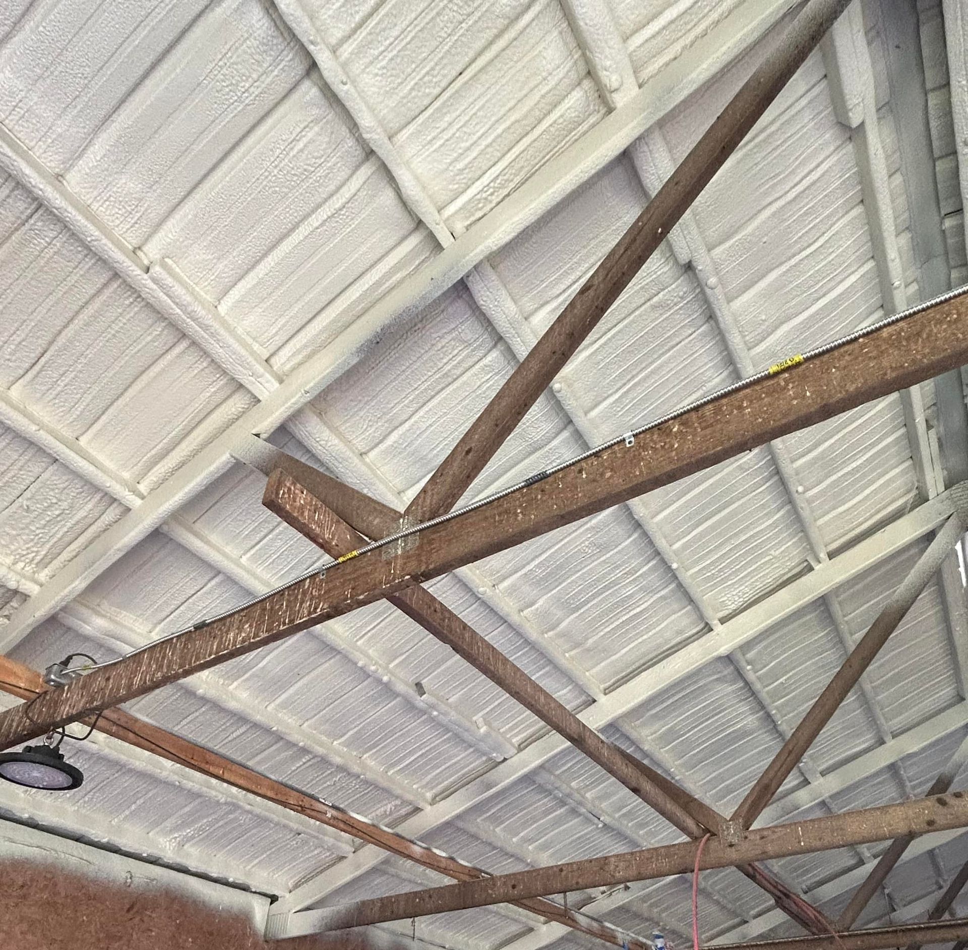 White spray foam insulation applied to the underside of a roof with exposed wooden beams and a light fixture.