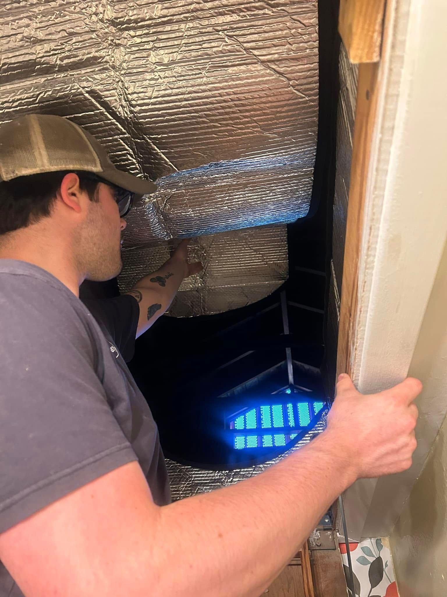 A man installs reflective insulation in a dark space. He wears a hat and glasses and holds a panel against a wall.