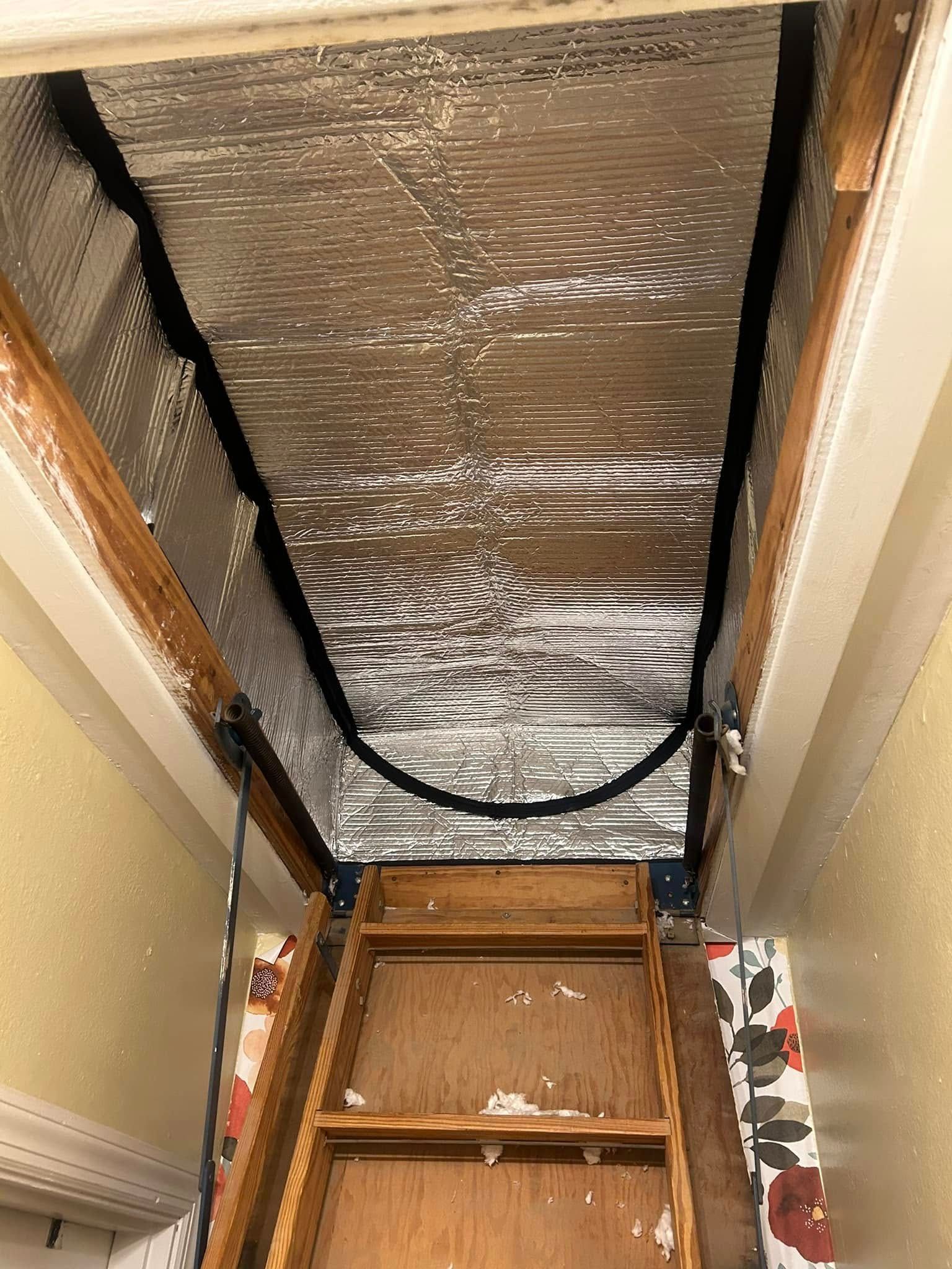 Attic access with pull-down wooden stairs, covered with reflective insulation. Wooden frame and floral wallpaper visible.