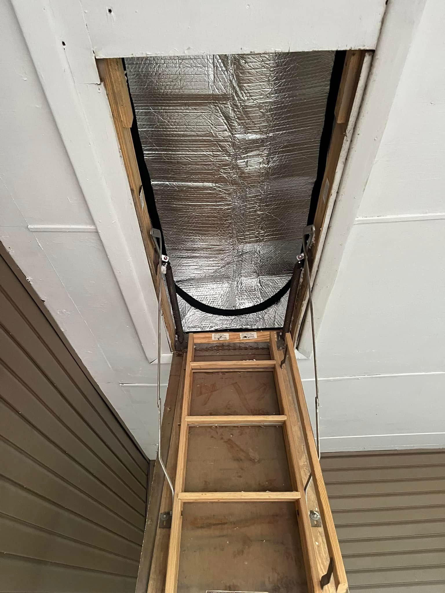 Wooden attic ladder extended downwards from an opening in a white ceiling. The opening is lined with reflective insulation.