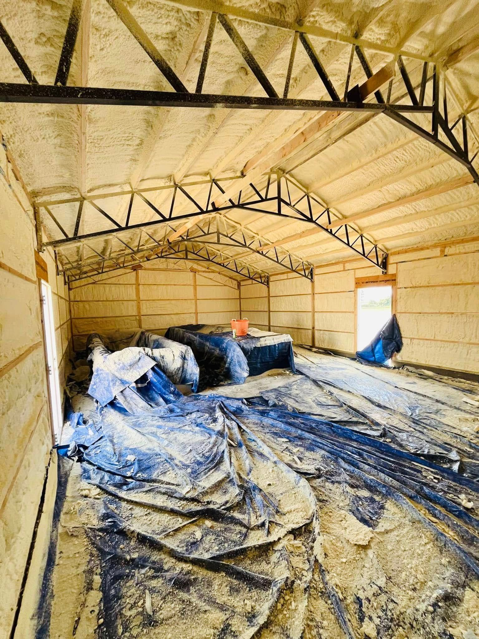 Interior view of a metal building with spray foam insulation. Blue tarps cover the floor, likely protecting contents.