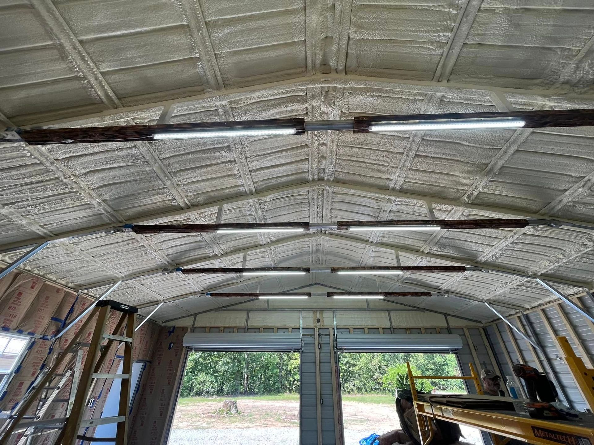 Interior view of a garage ceiling insulated with spray foam, featuring overhead lights and a partially open garage door.