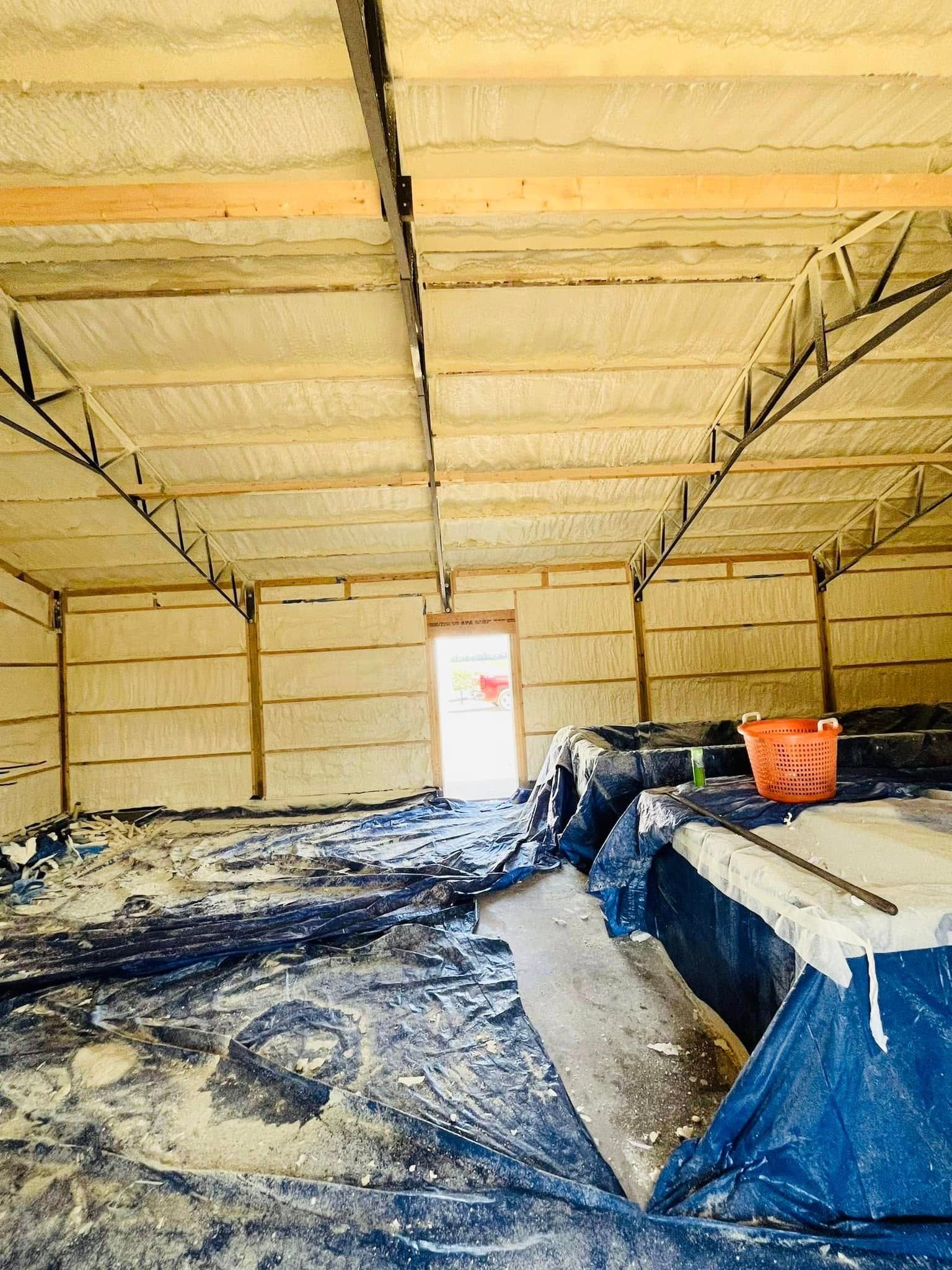 Interior of a building with spray foam insulation on walls and ceiling. Blue tarps and debris cover the floor, with an open doorway in the distance.