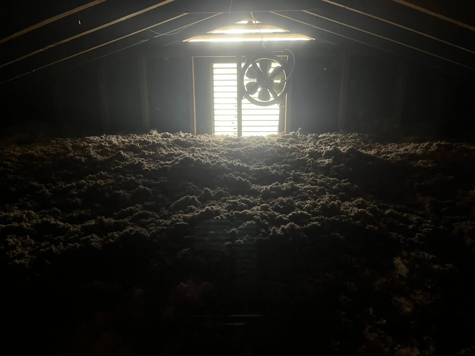 Dark, dusty attic with a thick layer of insulation. A bright window and fan are visible at the end, with light streaming in.
