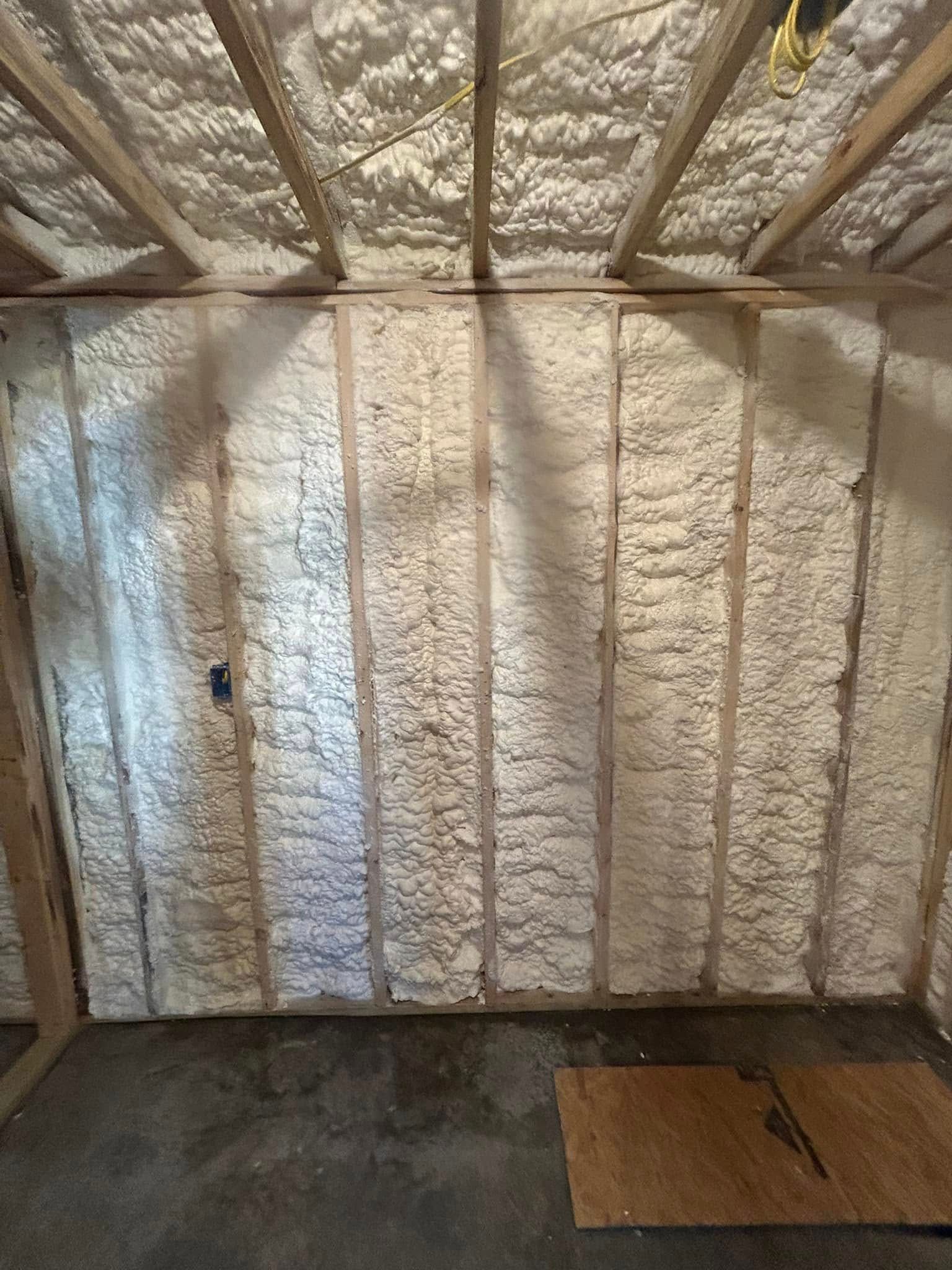 Interior view of a room with spray foam insulation covering the walls and ceiling, between wooden beams. The floor is concrete.