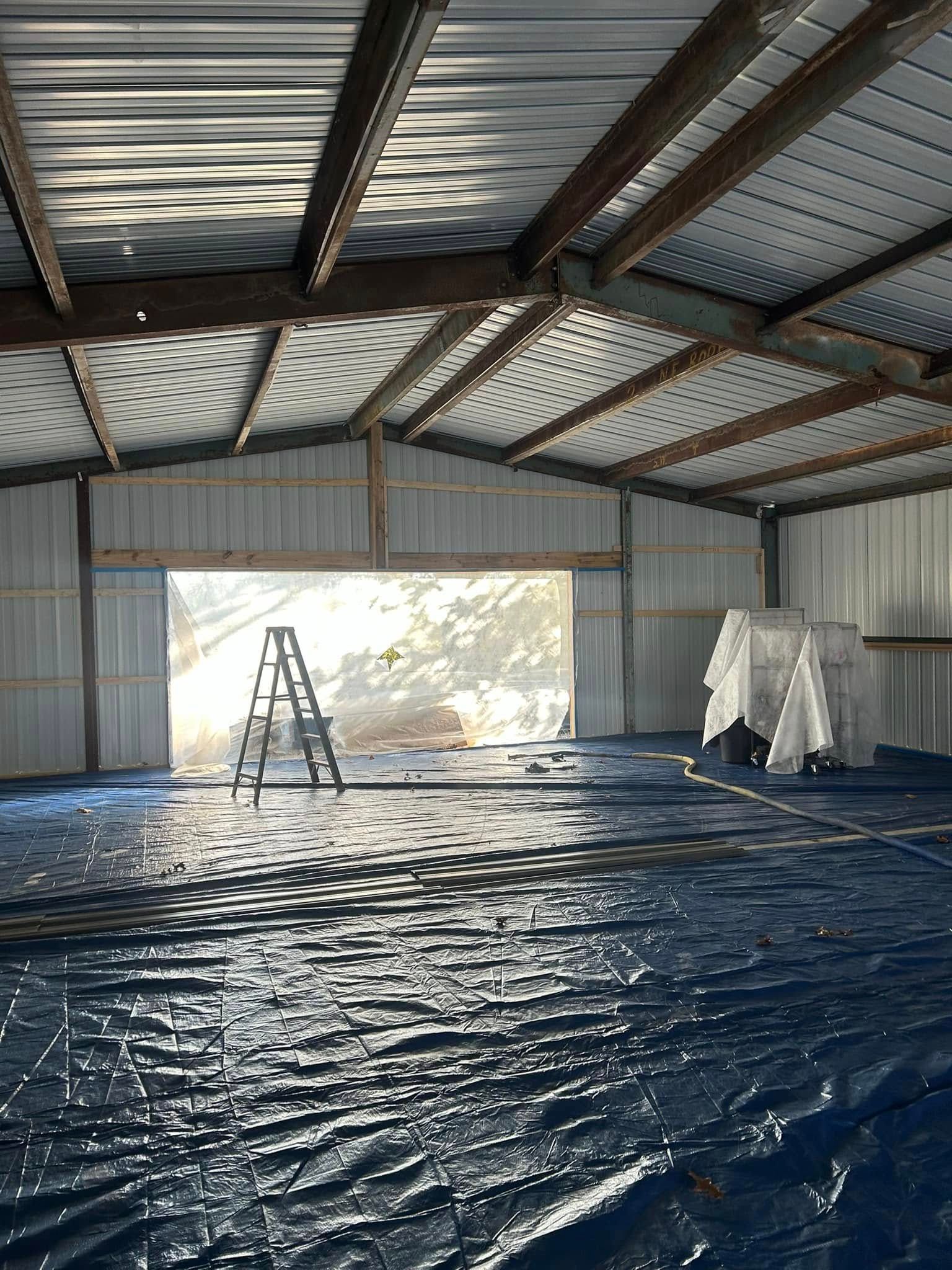 Interior of a metal building with a blue tarp covering the floor. A ladder stands in an open doorway.
